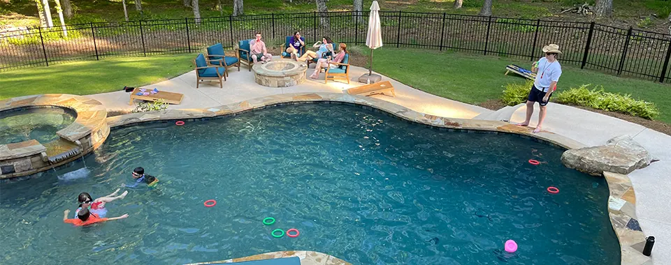 A large freeform pool seen from the deck, with a family seated around a fire pit and swimmers with pool rings.